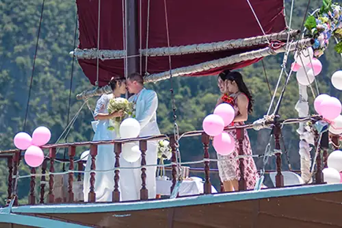 Just Married. A couple kiss as maids of honour look on, they are aboard a traditional junk style wooden boat decorated with flowers and pink and white balloons