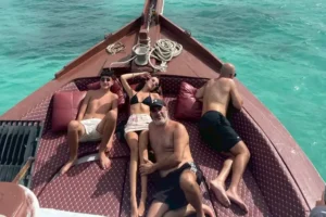 A family look to the camera as they lay on padded benches fitted into the bow of a wooden boat. They are dressed for a day of sunbathing at sea