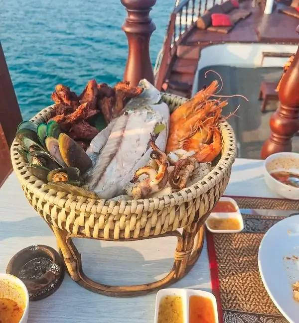 a basket of freshly barbecued seafood including, filleted fish, king prawns, squid, mussels and deep fried chicken. Served with dipping sauces. On a boat with the deck and the sea in the background.