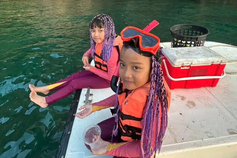 two young Asian children sit on the bow of a fibreglass boat. They are wearing lifejackets and snorkelling equipment, looking towards the camera