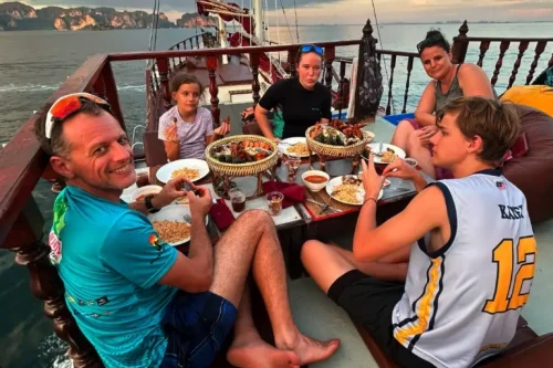 A family of five is gahtered around a low table on the high deck of a traditional wooden boat. They are dressed for warm weather, the sky looks the colour of sunset. They are eating a seafood BBQ buffet